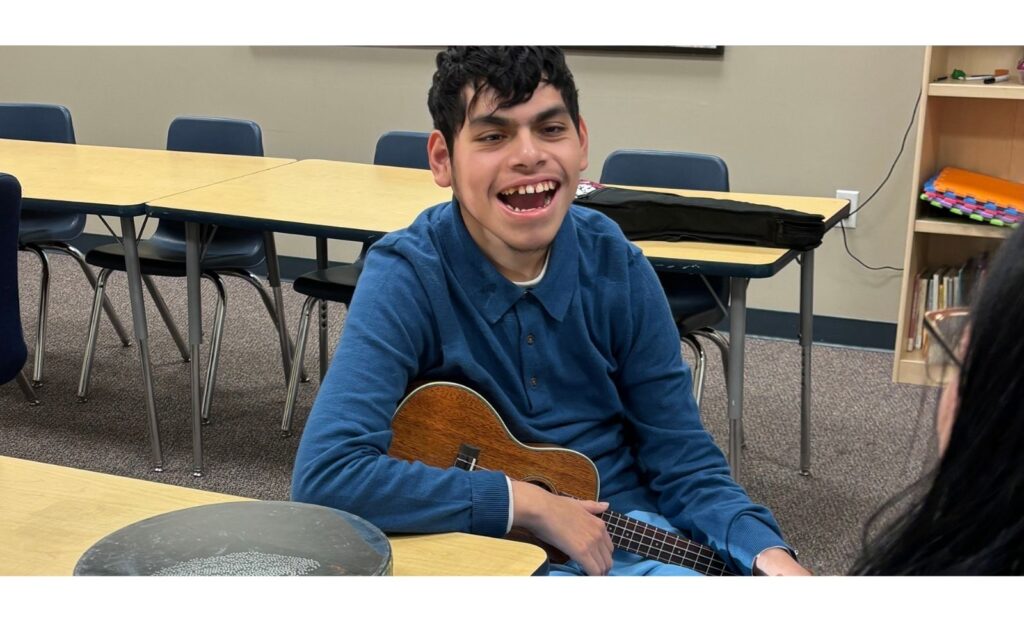 Smiling teenage boy with developmental disabilities holding a ukulele during a music therapy session in a classroom setting, engaging happily with the therapist
