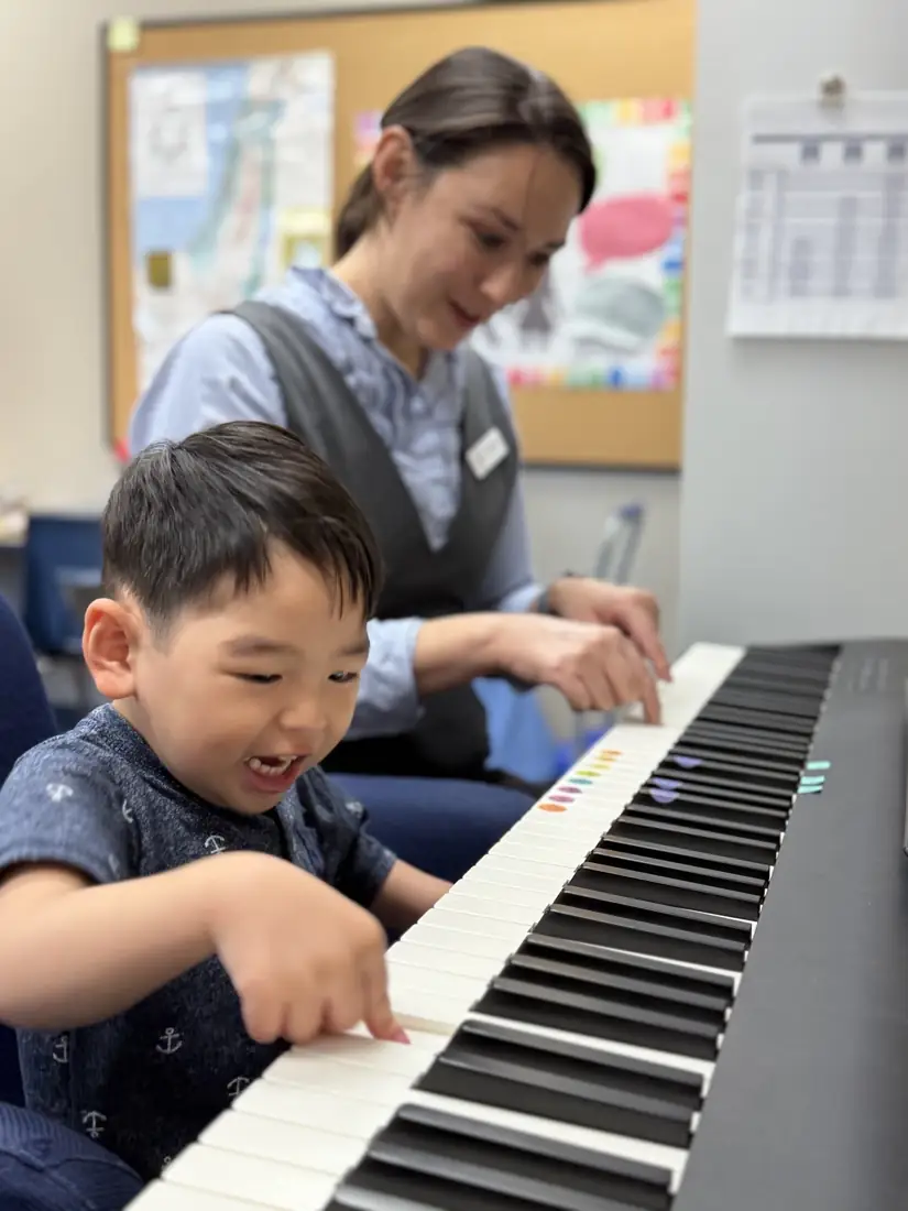 A young boy laughs as he plays the piano next to his smiling teacher