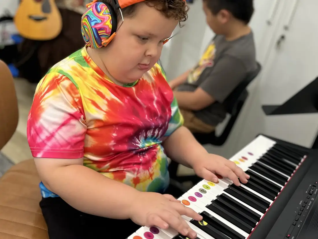 A young student in a rainbow tie-dye shirt and colorful headphones concentrates while playing a keyboard with color-coded keys