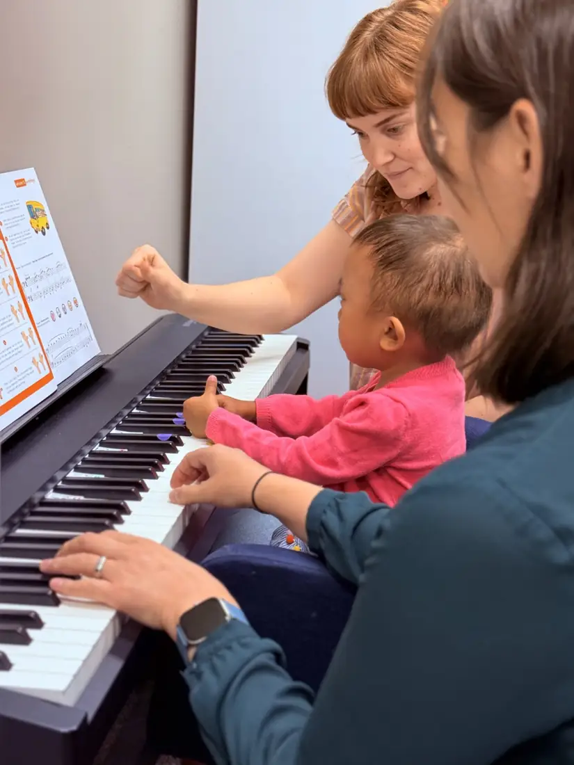 A teacher and parent cheer on a toddler in a pink sweater pressing the piano keys