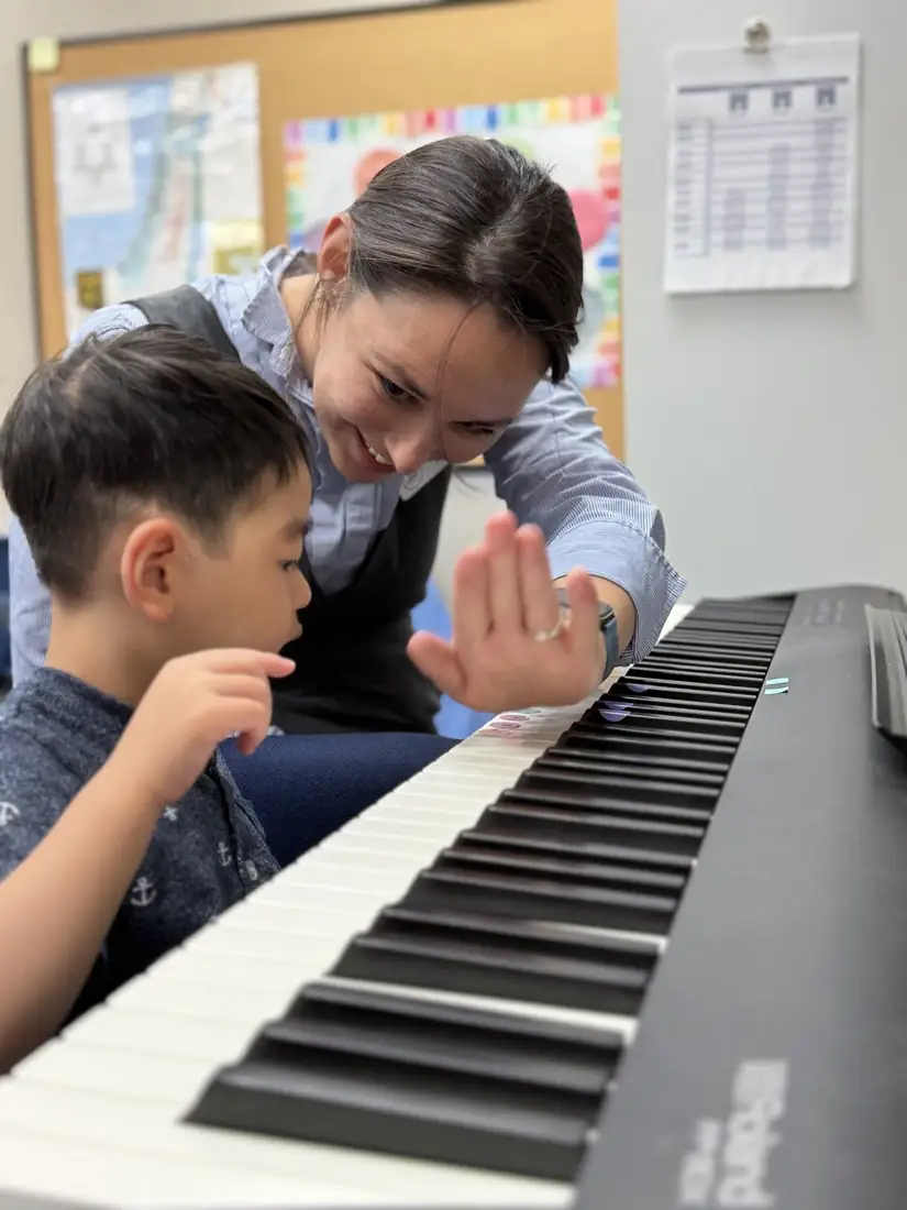 A smiling piano teacher holds up a hand for a high five as a young student focuses on the keys