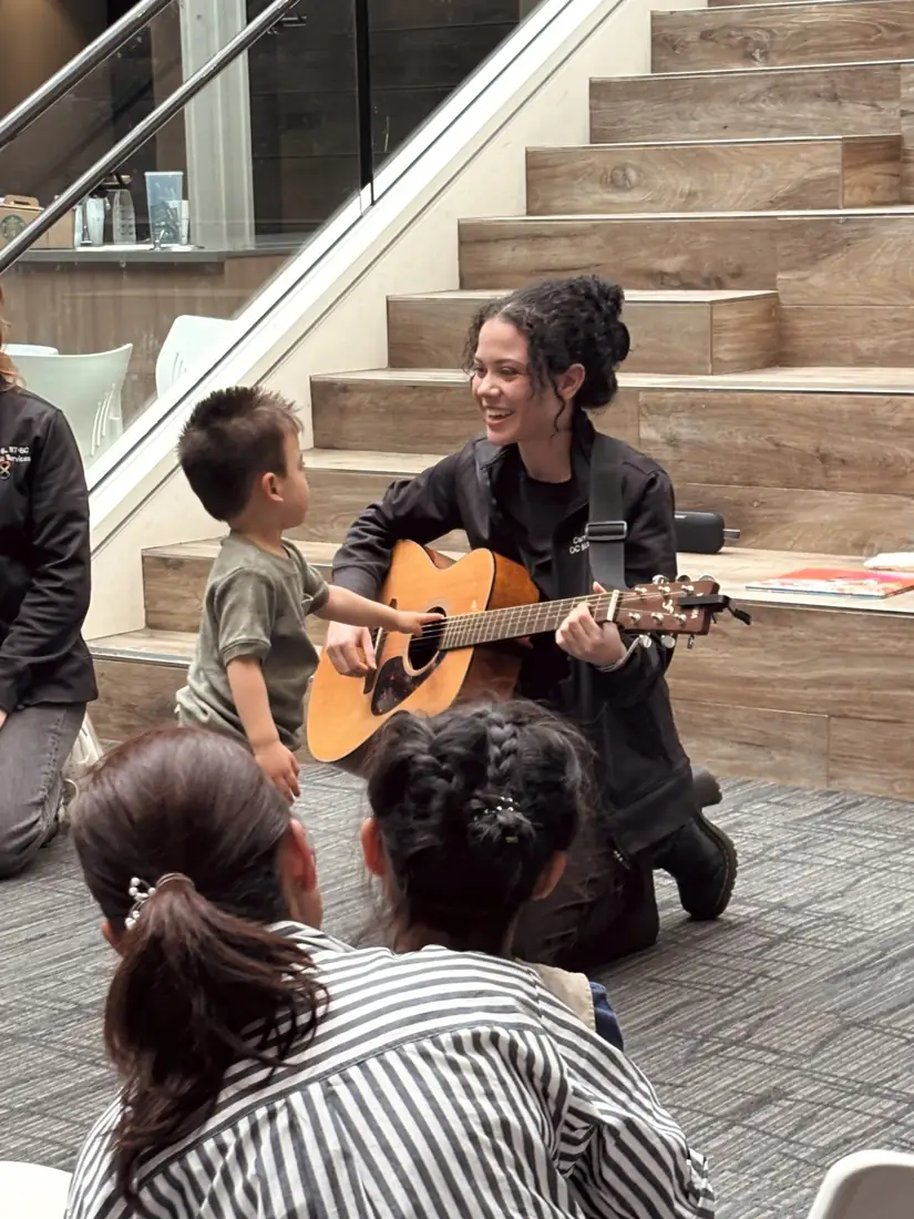A smiling teacher plays guitar while a toddler reaches out to strum