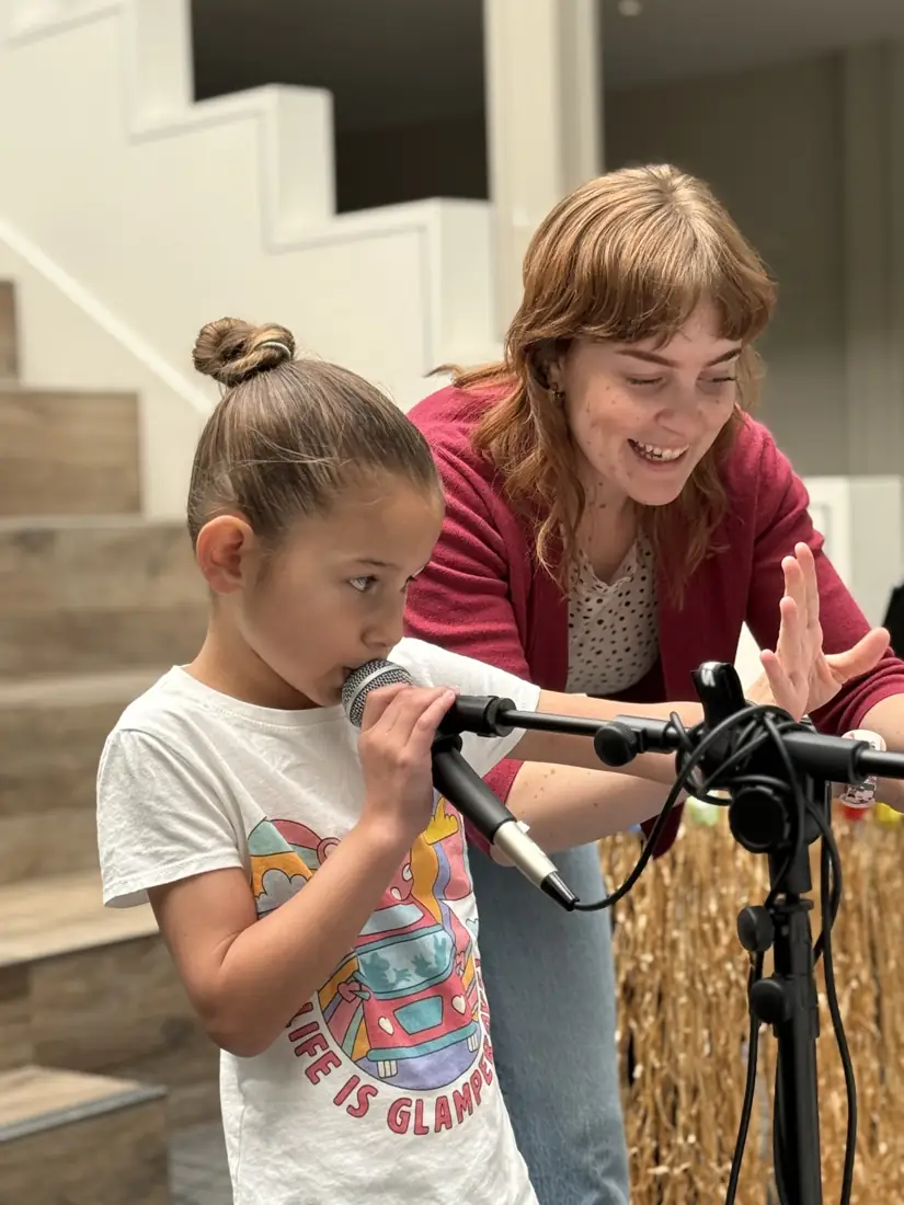 A young girl sings into a microphone while her smiling teacher cheers her on